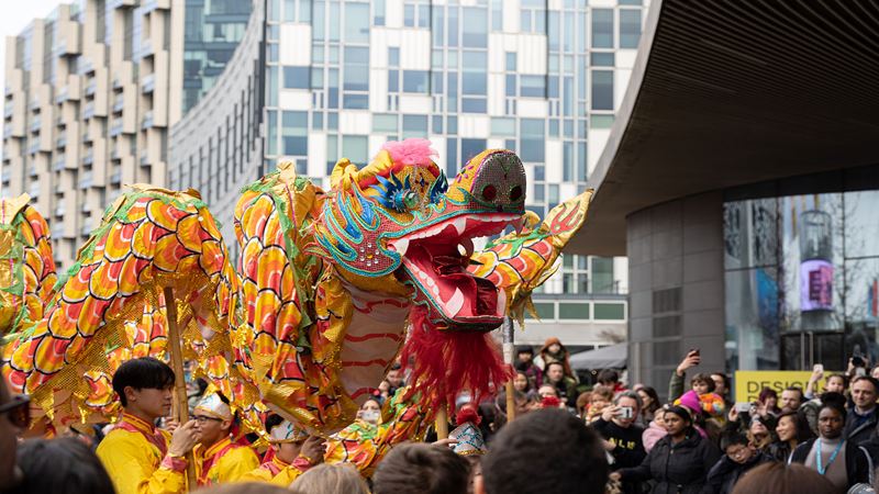 Dancers perform the Dragon Dance at Greenwich Peninsula surrounded by crowds of people and with big buildings in the background.
