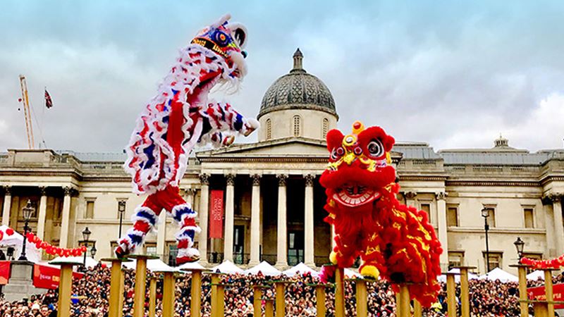 Two lion dance teams balance on yellow poles in Trafalgar Square, with crowds of people and the National Gallery behind.