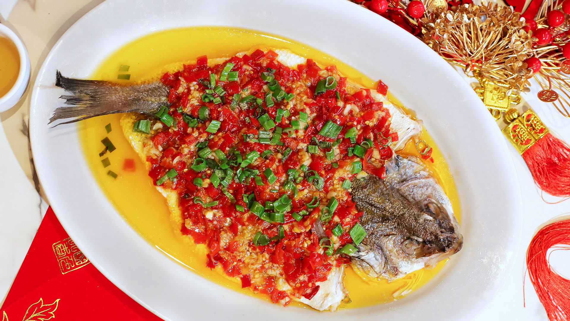 A plate of Turbot fish at China Modern. Image courtesy of Street & Co. Credit: Stacie Ma A close up of a whiite plate with a whole Turbot fish with green and red garnish on it on a decorated table.