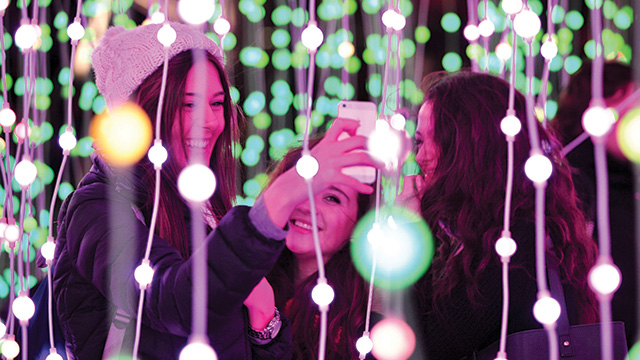 Christmas at Kew © RBG Kew. Image courtesy of Kew Gardens. Three women take a selfie among strings of green and white fairy lights at night.