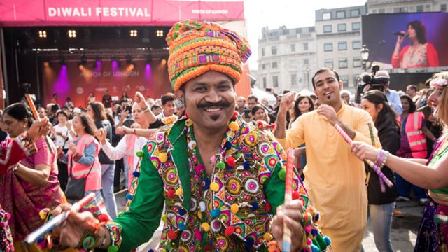 Diwali on Trafalgar Square. Credit: Greater London Authority. Image courtesy of the Greater London Authority. A man dressed in traditional clothing with flower garlands around his neck, at the Diwali festival, while other performers dance in the background.