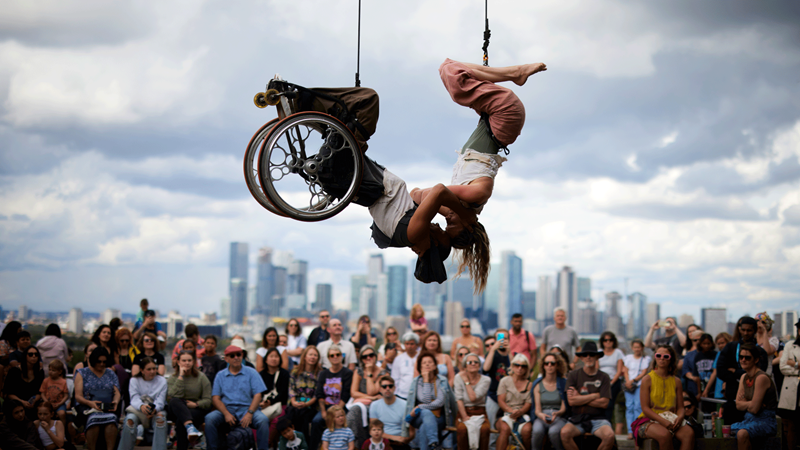 Be amazed by the lineup of showstopping live events at the free Greenwich+London Docklands International Festival. Credit: David Levene. Two performers hang from ropes during a performance in front of crowds with the City of London in the background