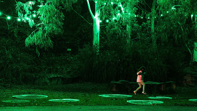 A girl jumps on giant lily pads illuminated in green as trees behind are lit with green lighting.