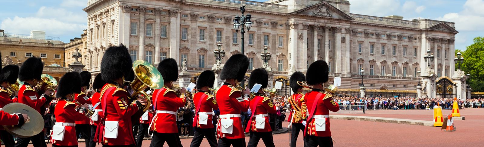 The kings guard in red uniform with large black hats marching past crowds outside buckingham palace.