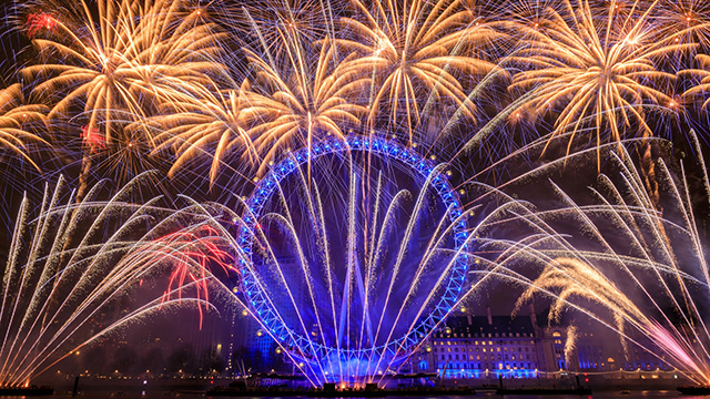 The London Eye at night illuminated in blue, with blue, yellow and red fireworks exploding around it.