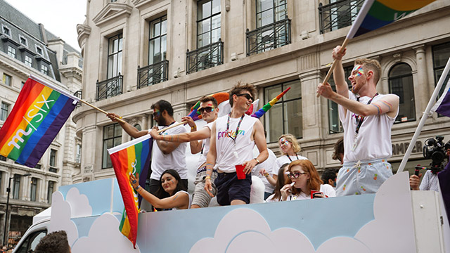 Pride in London. Credit: @lifeofsiobhan. Image courtesy of @lifeofsiobhan. People standing up on a float, waving rainbow flags, as they travel along during the Pride in London parade.