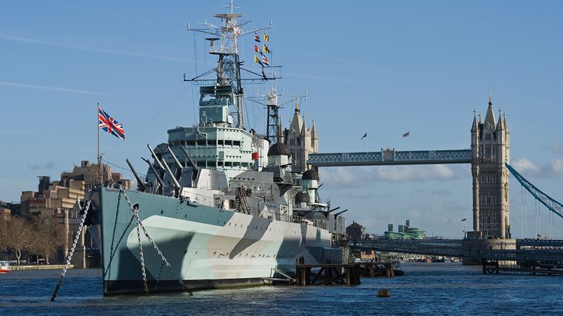 Take a tour of the famed HMS Belfast on the river Thames. Credit: Richard Ash. Image courtesy of IWM. A battleship sits atop the water with a bridge and blue sky in the background.
