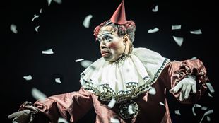 Billy Porter plays The Emcee in the musical Cabaret at the Kit Kat Club, wearing a red clown outfit with white confetti flying around.