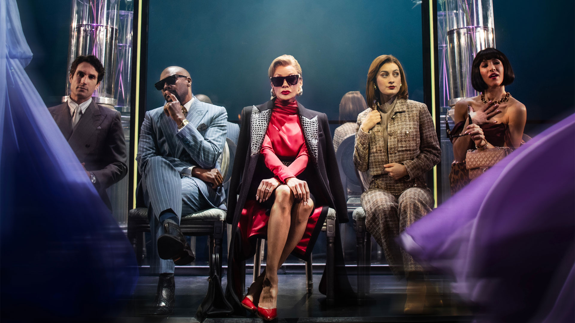 Relive high-stakes fashion moments when seeing London’s new musical The Devil Wears Prada. Photo credit: London Theatre Direct. Actress Vanessa Williams, playing Miranda Priestley in the musical The Devil Wears Prada, is seated centre stage in a red dress and sunglasses, staring at the audience and surrounded by colleagues.