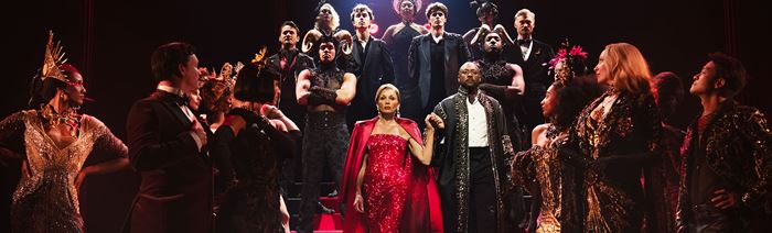 Miranda Priestly, played by Vanessa Williams, wears a glittering red dress and walks down the stairs surrounded by the cast of Devil Wears Prada.