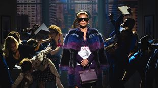 Vanessa Williams, portraying editor-in-chief Amanda Priesley, wears black sunglasses, a purple cape and a handbag as she walks across the stage while the press ask her questions.