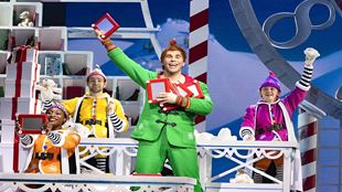 The actor playing Buddy in ELF the Musical is wearing a flashy green uniform and holding some toys and surrounded by Santa's little helpers all dressed with colourful costumes.