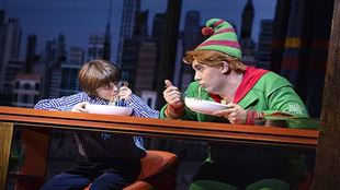 Buddy, wearing a red ad green elf costume stands next to his half brother eating a bowl of pasta.