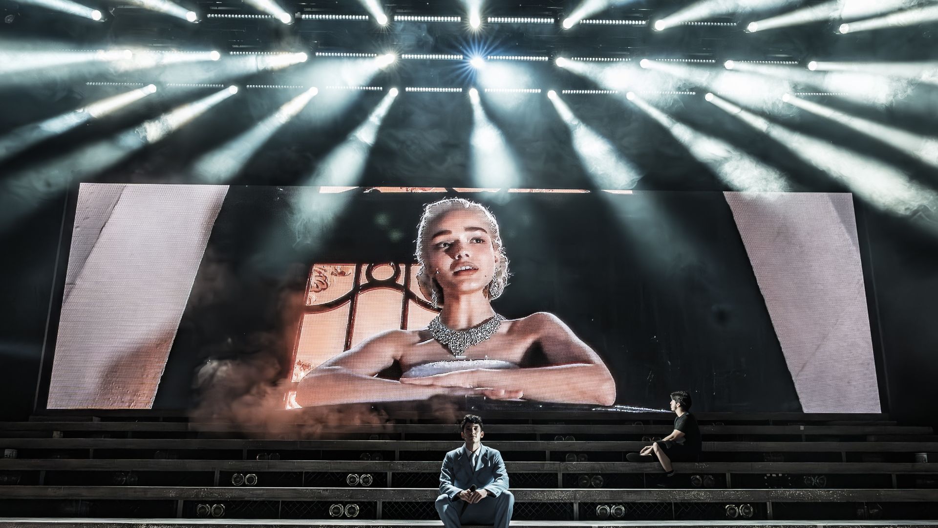 See Rachel Zegler star in the revival of Andrew Lloyd Webber and Tim Rice's legendary musical Evita at the London Coliseum. Image courtesy of London Theatre Direct. large tiered west end stage with an actor sat at the bottom of the tiered steps, and another actor sat a few rows behind looking up at a screen with a woman performing a song