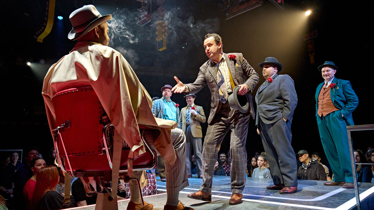 See Guys and Dolls at the Bridge Theatre and follow Nathan Detroit in the streets of Manhattan as he gambles his way out of a police investigation. Image courtesy of Premier Entertainment/ photo credit Manuel Harlan. Several men in three-piece suits are standing on the stage of Guys and Dolls, facing another man sitting in a chair.