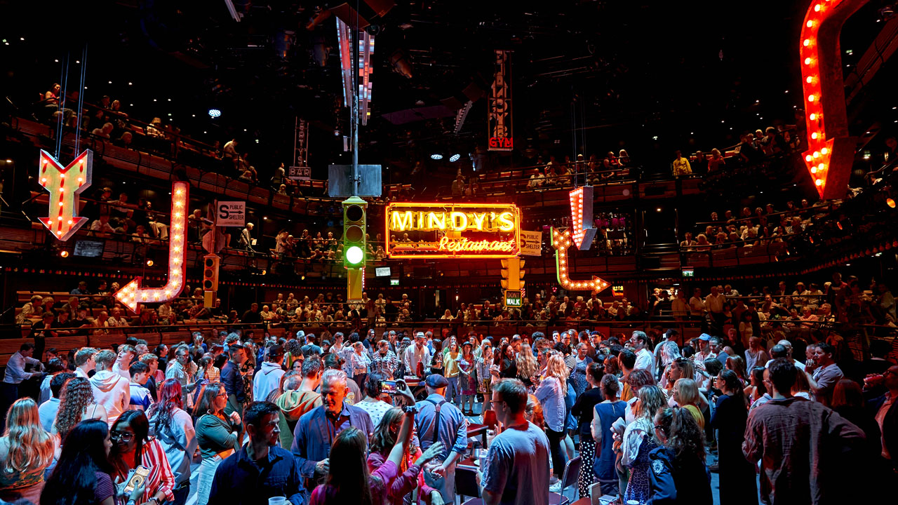 See the Broadway classic Guys & Dolls take over the London West End at the Bridge Theatre. Image courtesy of Premier Entertainment/ photo credit Manuel Harlan. The Bridge Theatre is crowded with the audience surrounding the stage of Guys & Dolls and its big Neon lit sign.