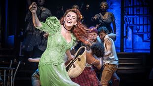The actress and Hadestown performer Rachel Tucker iswearing a long light green dress and singing on the stage of the Lyric Theatre. 