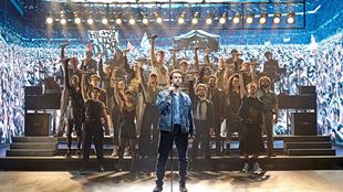 A man stands at the front and sings while back-up singers stand at the back, all raising their hands to the sky during a performance of Just For One Day - The Live Aid Musical. 