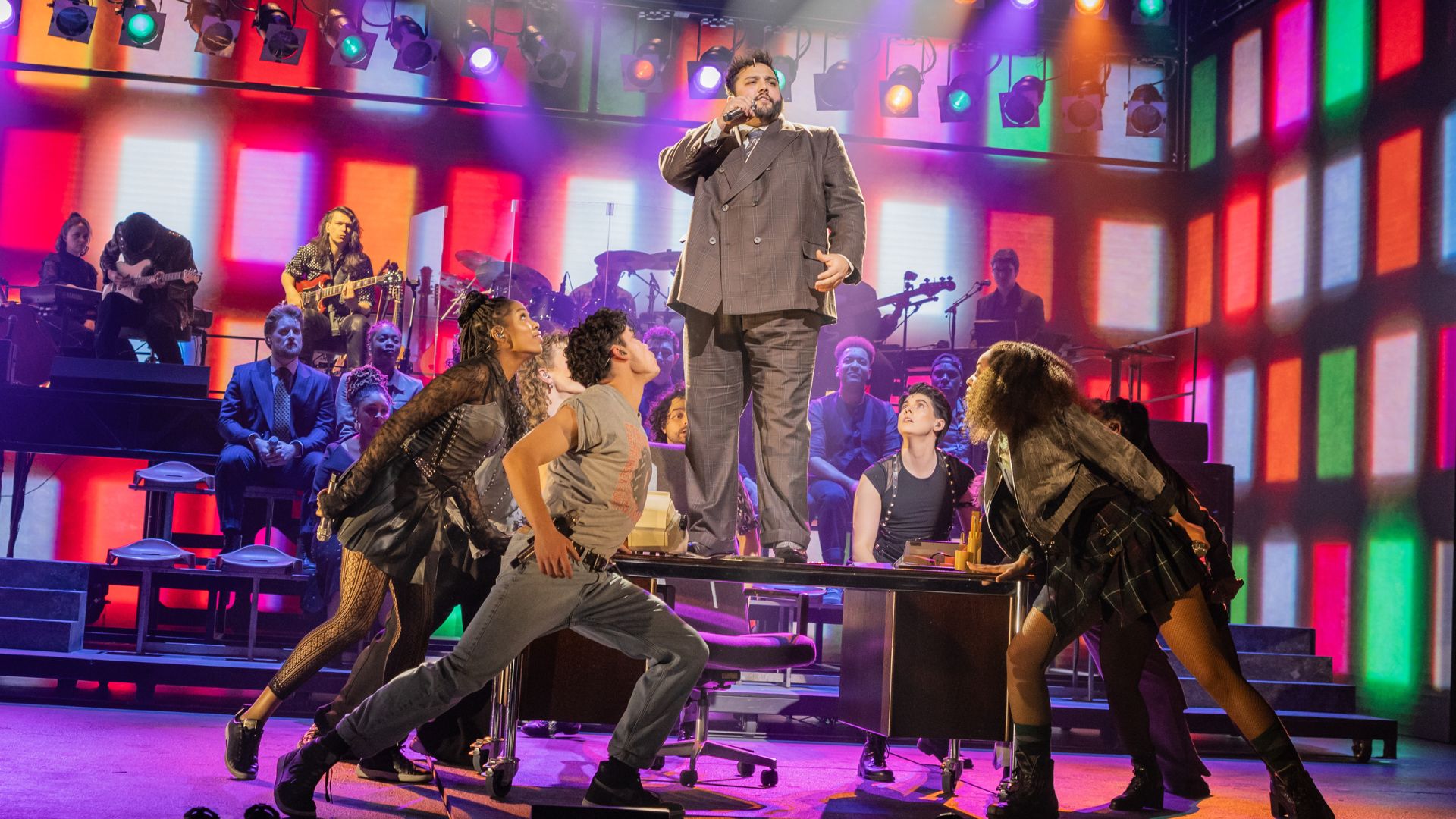 Celebrate the 40th anniversary of the gig that rocked the world with the new musical Just For One Day – The Live Aid Musical at the Shaftesbury Theatre. Image courtesy of London Theatre Direct. The cast of Just For One Day stand around a performer stood singing on a table and perform a song, with a screen in the background showing bright 80s colours.