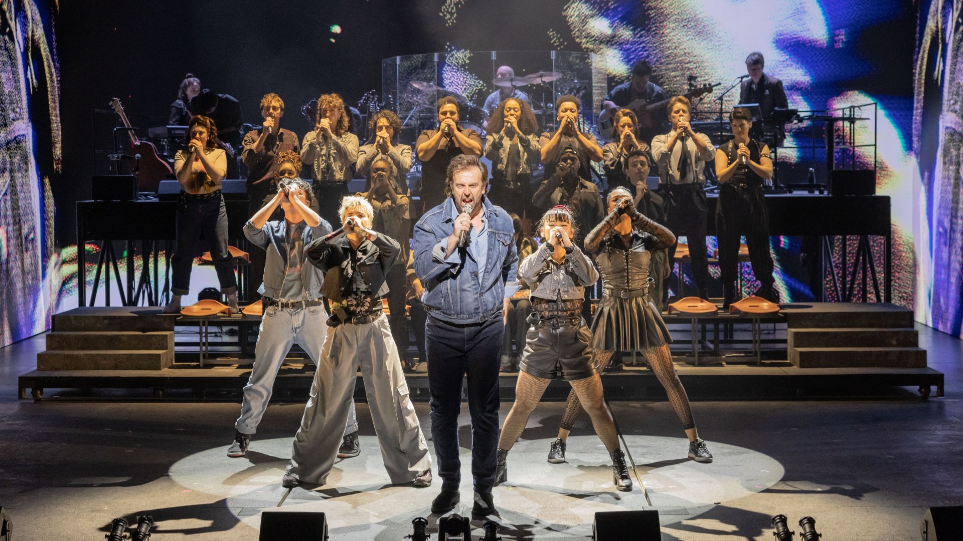 Relive the sensational concert that captured the world's attention with top rock stars in the new musical Just For One Day at the Shaftesbury Theatre. Image courtesy of London Theatre Direct The cast of Just For One Day stand in the centre of the stage and perform a song, with a screen in the background and a live band.