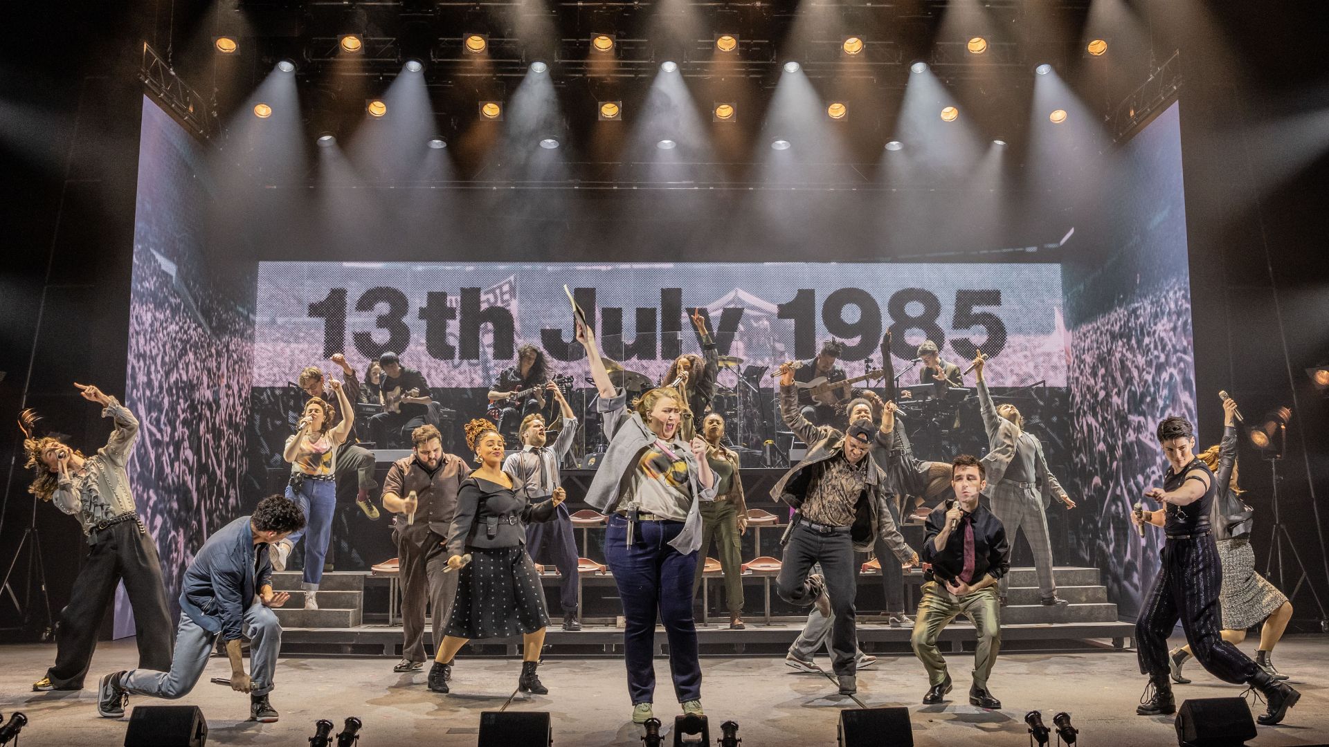 Hear hits from historic bands and singers including Queen and Bowie in the London musical Just For One Day – The Live Aid Musical. Image courtesy of London Theatre Direct The cast of Just For One Day fill the stage and perform a song, with a screen in the background showing footage from the Live Aid concert.