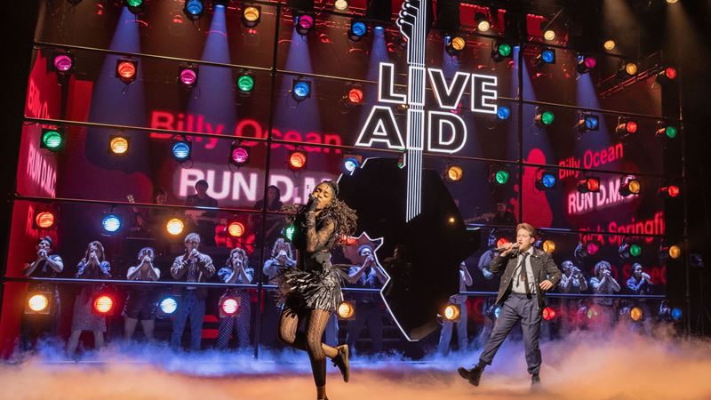 The cast of Just For One Day perform a song with a screen in the background showing lights and the live aid guitar symbol.