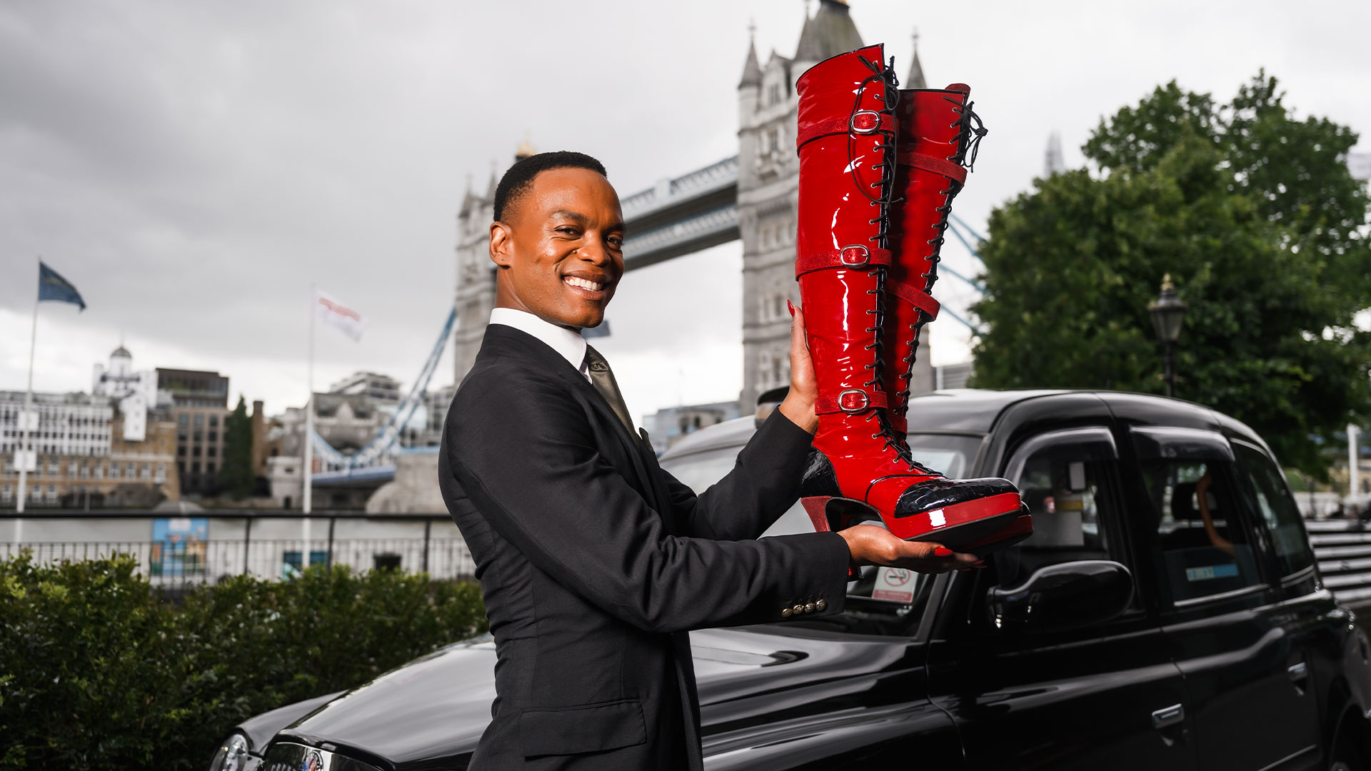 See the return of Kinky Boots to the London West End with Johannes Radebe to reprise the iconic role of Lola. Photo credit: Charlie Flint. Against a black-and-white backdrop featuring a black cab and Tower Bridge, Johannes Radebe holds the iconic pair of red high-heeled boots.