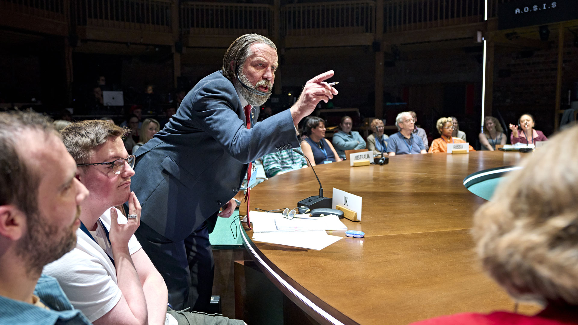 Plunge into the heart of the 1997 UN Climate Change Conference with the gripping play Kyoto at London's Soho Place Theatre. Image courtesy of RSC & Good Chance/ Photo by Manuel Harlan. A man wearing a dark grey suit, representing the United Kingdom in the play The Kyoto, stands and points his finger at someone on stage.
