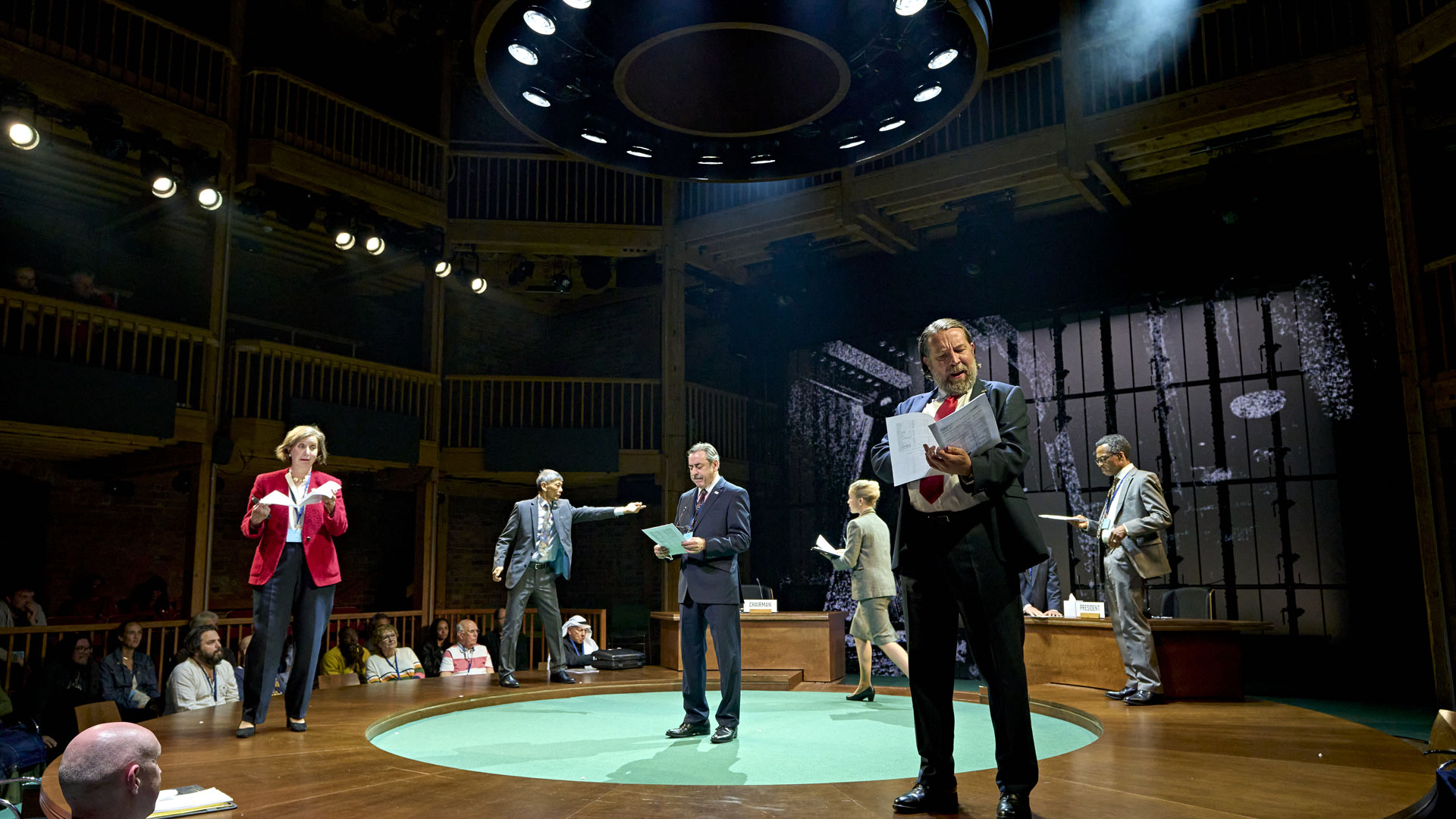 Watch key nations and UN officials debate climate change in the play Kyoto, coming to London's West End in January 2025. Image courtesy of RSC & Good Chance/ Photo by Manuel Harlan. Actors stand on a circular stage holding policy documents and debating during the production of the play Kyoto.