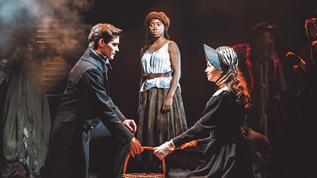 A man and a woman in period costume, looking at each other while holding a basket while a woman standing behind looks at them on a dark evening.