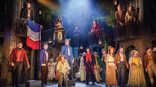 The cast of Les Misérables, dressed in period costume, stand on the stage facing the audience, with a French flag in the background. 