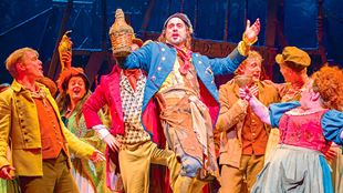  A man wearing long blue and brown trousers in a military style is holding a bottle and is surrounded by other members of the cast of Les Misérables.