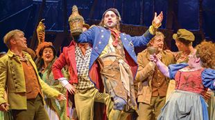 A man wearing long blue and brown trousers in a military style is holding a bottle and is surrounded by other members of the cast of Les Misérables.