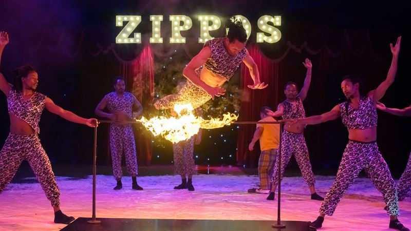 Admire the Timbuktu Troupe perform with the Zippos Circus, a spectacular showcase of acrobatics and firebreathing excellence. Image courtesy of Zippos Circus Nomads 2023/ Photographer: Piet-Hein Out A man jumps above flames set up by team mates surrounding him on the stage of Zippos Circus.