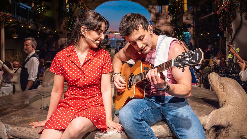 Immerse yourself in a story of family tradition and passionate, budding love with Mamma Mia! The Party. Credit: About Grace PR. Image courtesy of About Grace PR. A woman wearing a red polka dot dress is sitting next to a man playing guitar at Mamma Mia! The Party.