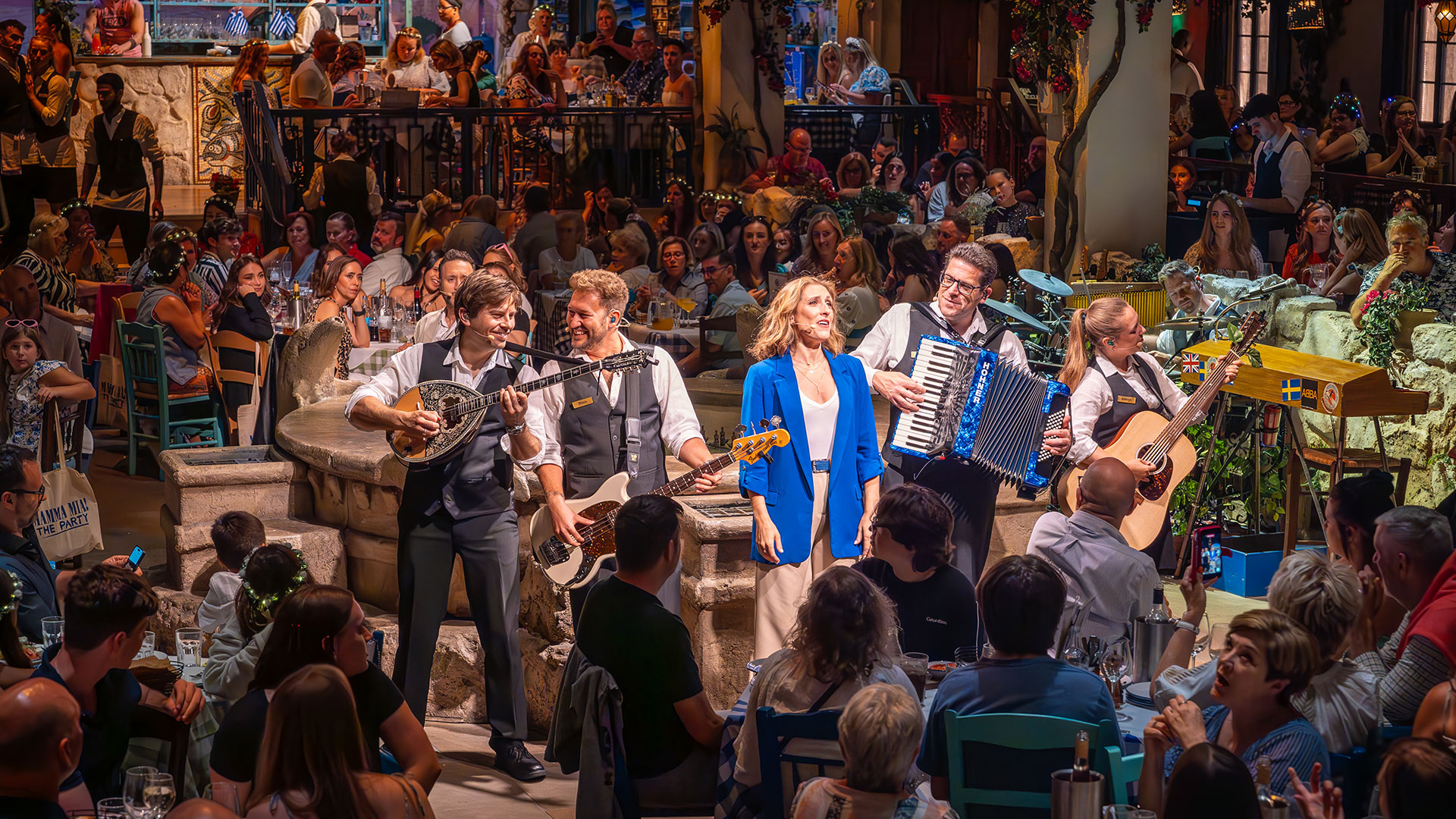 A look inside Mamma Mia! The Party at The O2. Image courtesy of Marc Brenner. A woman in a blue jacket sings, accompanied by a band, surrounded by an audience seated at tables around the room at Mamma Mia! The Party.
