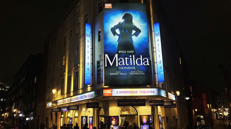 Outside the Cambridge Theatre, where theatregoers are waiting for the evening performance of Matilda. The theatre has a giant poster of the show, which is illuminated at night.