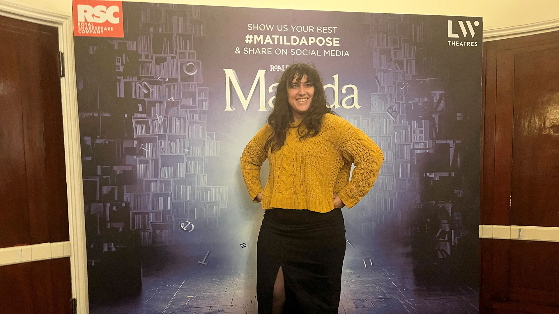 Channel your inner Matilda and strike the iconic pose during the interval of Matilda the Musical at the Cambridge Theatre. © Carole Rocton A woman in a bright yellow sweater and long black shirt stands in front of the Matilda the Musical poster, striking the iconic power pose.