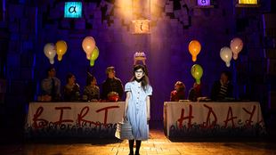 The actress playing Matilda stands at the centre of the stage, wearing a light blue dress, while other children sit around a table with balloons and a tablecloth that spells out "BIRTHDAY" in bright, bold red letters.