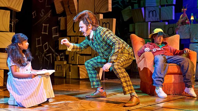 A man wearing a tartan two piece suit seems to be arguing with a young girl sitting on a pile of books.
