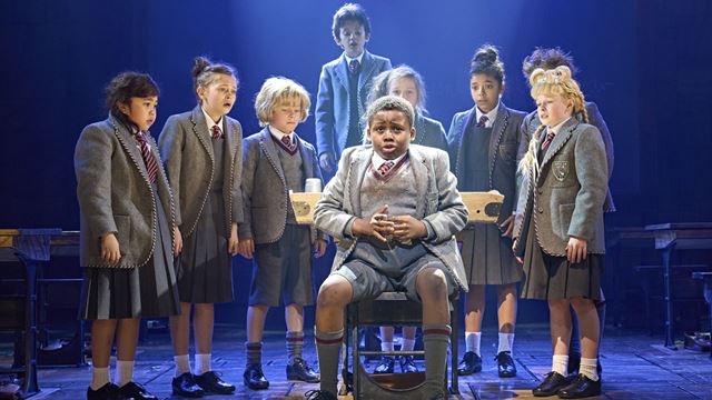 Matilda The Musical performs on the London West End. Image courtesy of See Tickets. A group of school kids are surrounding one of their classmates seating on a chair during the show Matilda The Musical.