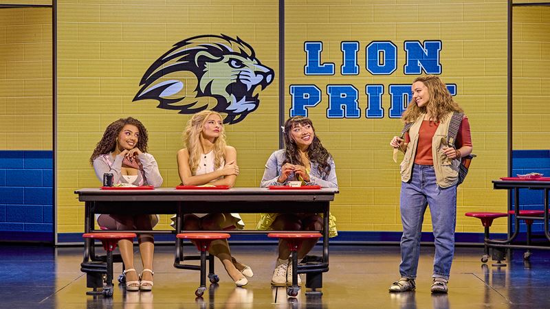A staged classroom with three teenage girls that sit at a table and one other girl standing next to them all look at each other with a lion head symbol and the blue text lion pride on a yellow wall. 