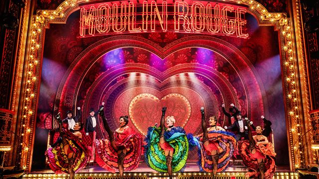 A troup of dancers wearing colourful dresses are dancing the French Cancan on the stage of Moulin Rouge The Musical in London.