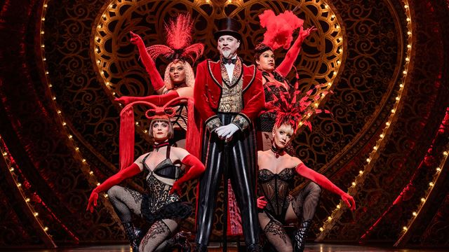 Harold Zidler, the owner of Moulin Rouge, stands on the front stage of the Picadilly Theatre with four cabaret dancers standing behind him dressed with lace corsets.
