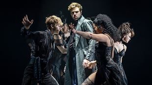 In the musical Moulin Rouge, a man dressed in a long light blue coat is surrounded by dancers dressed in black.