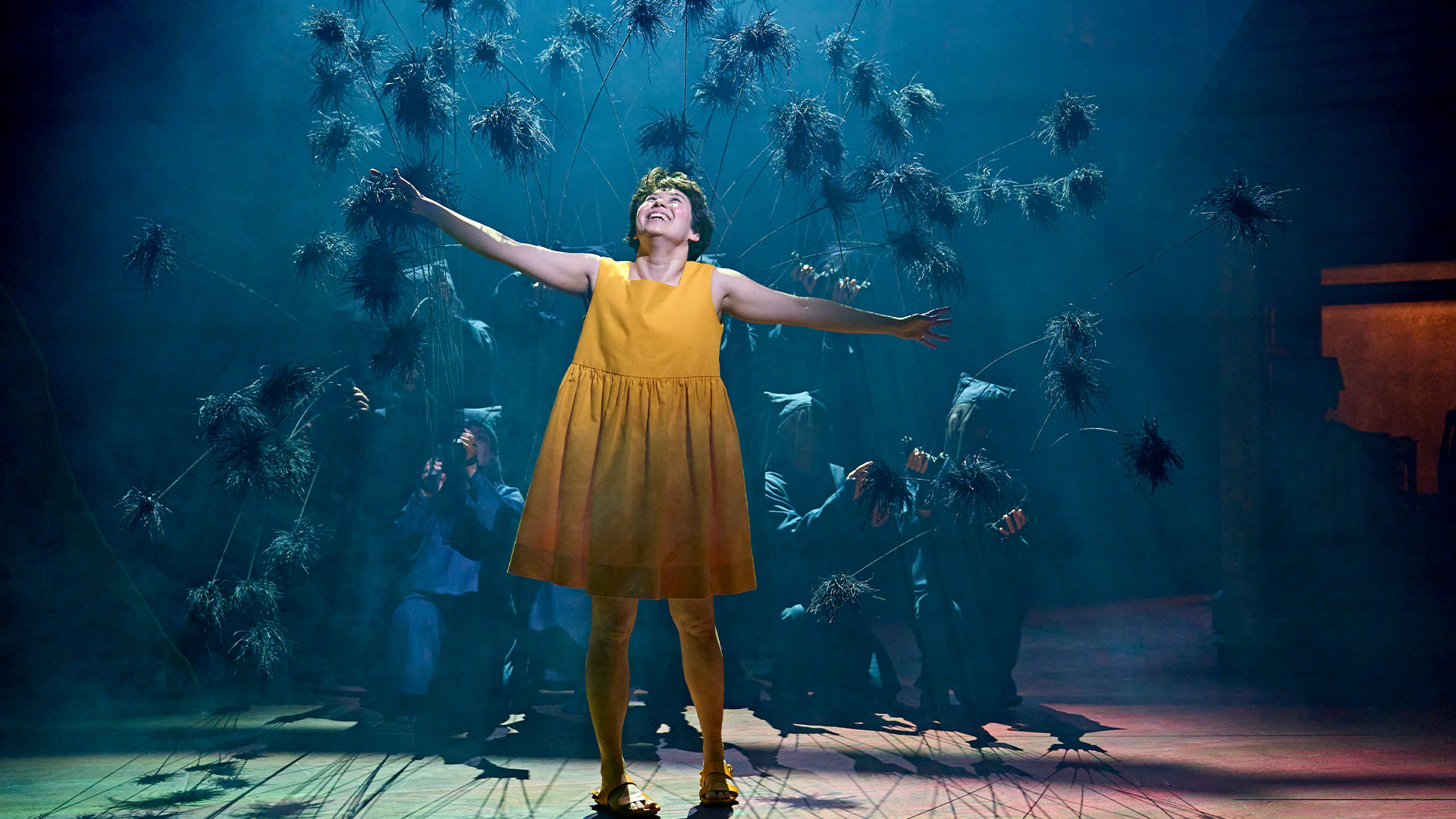 Discover the story of two sisters exploring the unseen world of forest spirits in the stage adaptation of the world-famous Ghibli anime, My Neighbour Totoro. © Manuel Harlan Satsuki (Ami Okumura Jones) wearing a yellow dress and holding her arms out as the Soot Sprites go around and behind her.