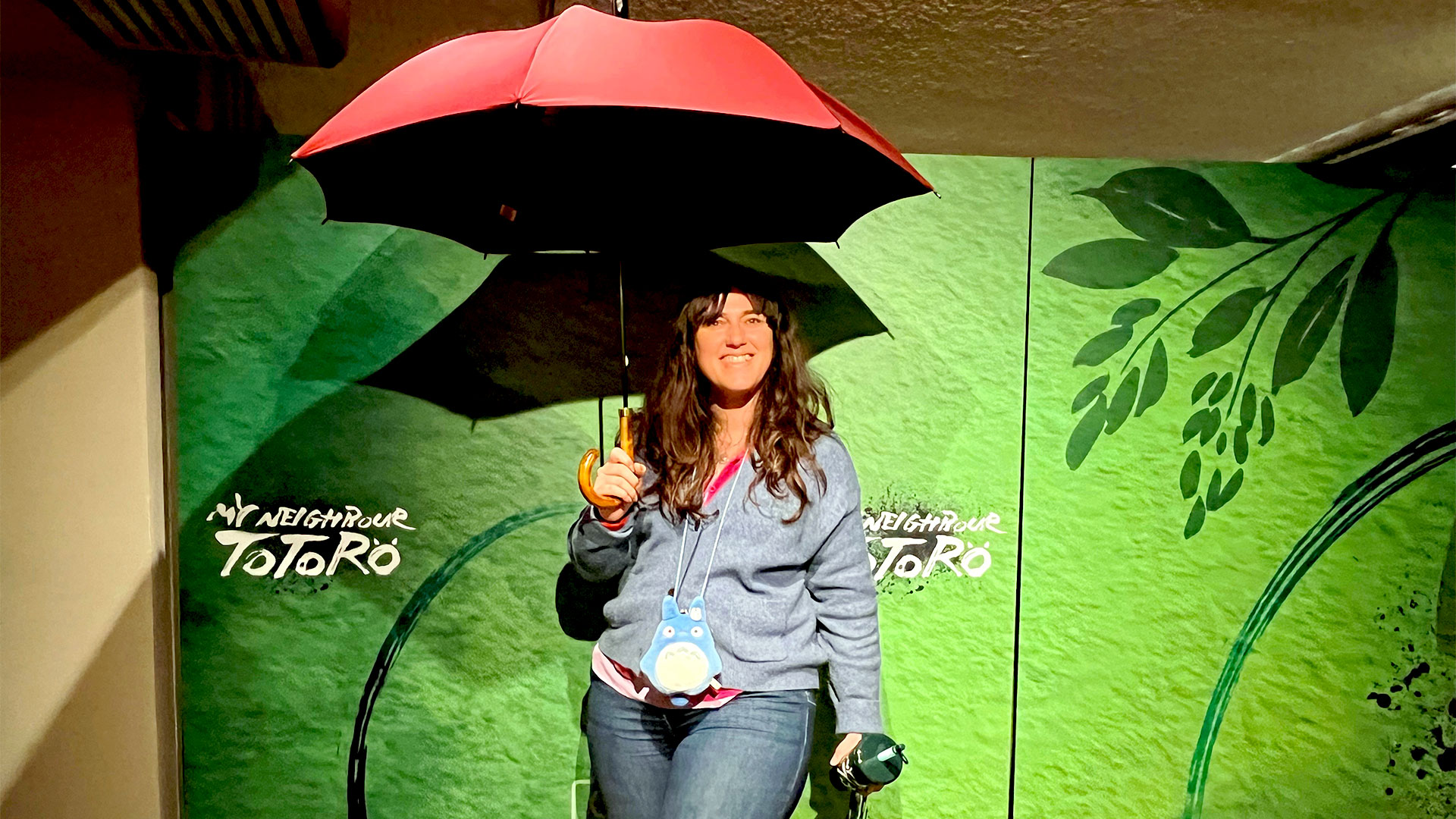Make some unforgettable memories by going to see the Olivier Award-winning play My Neighbour Totoro. Photo credit: Carole Rocton. A woman stands under a red umbrella against a green backdrop, which is part of the stage adaptation of My Neighbour Totoro at the Gillian Lynne Theatre.