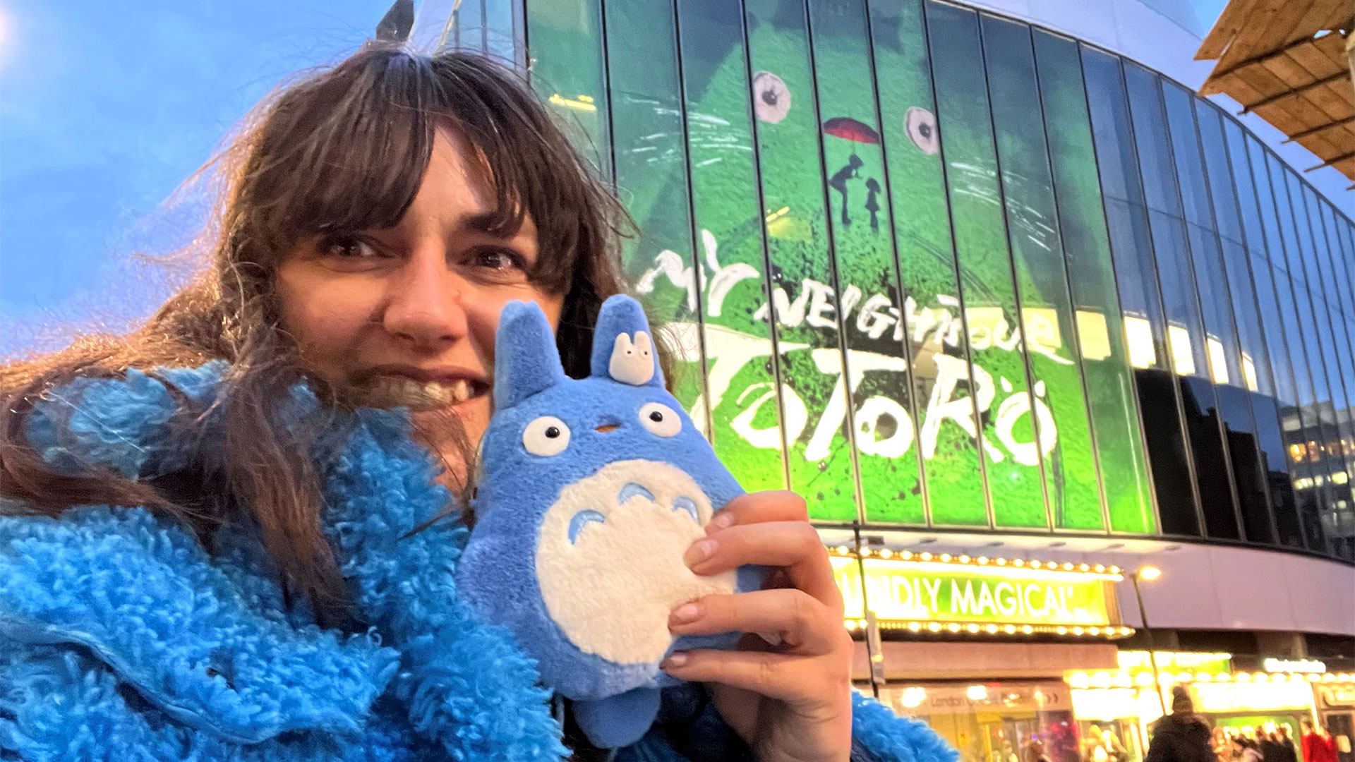 Head to Covent Garden to see the sensational stage production of My Neighbour Totoro, currently showing in London's West End. Photo credit: Carole Rocton. A woman wearing a bright blue, furry coat is standing in front of the Gillian Lynne Theatre, holding a blue Totoro plush toy.
