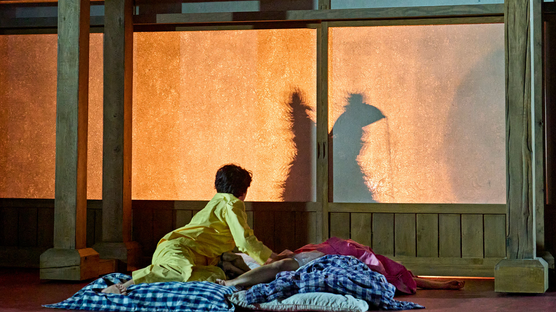 Step back into your childhood and rejoice as Studio Ghibli's fan favourite, Totoro, takes to the stage at the Gillian Lynne Theatre. © Manuel Harlan Satsuki (Ami Okumura Jones) wearing yellow pajamas and looking at a shadow of Totoro as Mei (Victoria Chen) sleeps next to her.