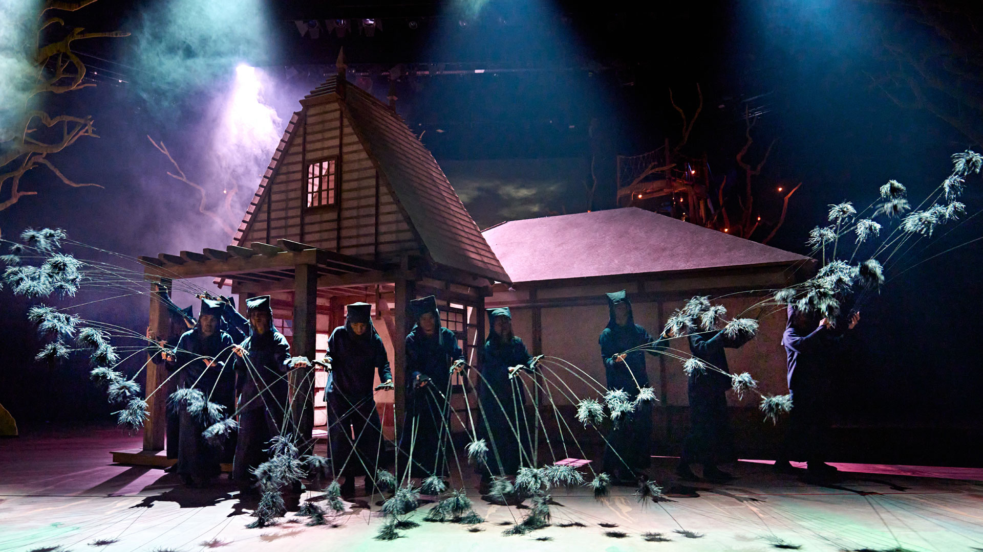 See the whimsical spirit of the forest return to the stage of the Gillian Lynne Theatre in the incomparable My Neighbour Totoro. My Neighbour Totoro production images, 2023. Photo by Manuel Harlan (c) RSC, with Nippon TV Members of the cast of My Neighbour Totoro direct the puppets portraying the little black sooty creatures surrounding a house on stage.